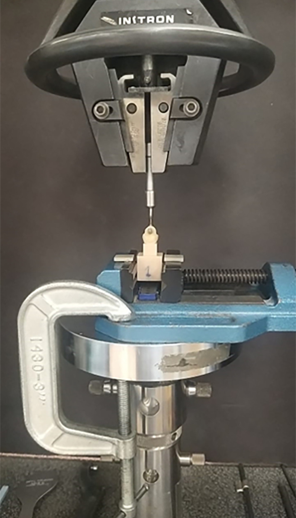 The universal testing machine demonstrates the connection of the test samples for the pull-off test, where the handles of the zirconia crowns are positioned to align with the sample handle in a perpendicular manner to the direction of the pulling force. The image was captured using a Canon EOS 700D digital single-lens reflex camera equipped with a 100-mm macro lens, either with or without a ring flash.